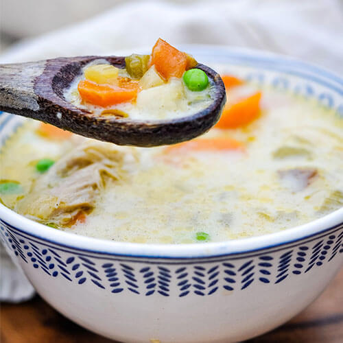 wooden spoon scooping into a colorful veggie soup of chicken, carrots, and peas