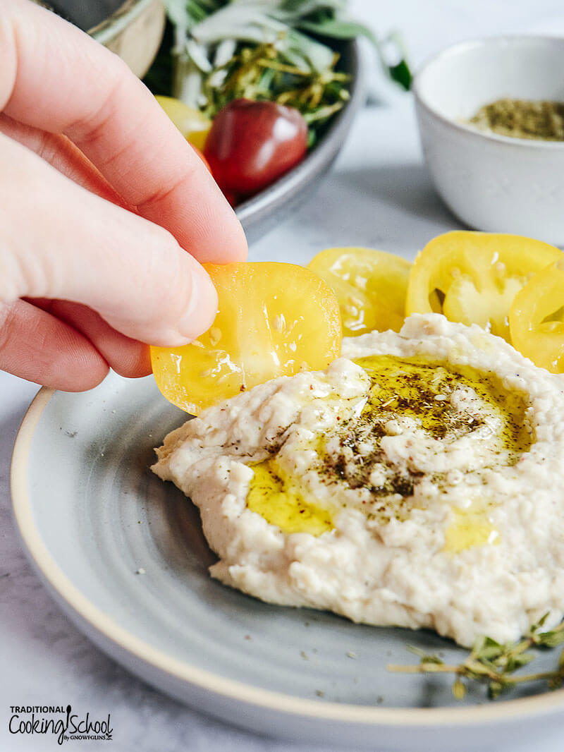 woman's hand scooping up homemade hummus with a tomato slice
