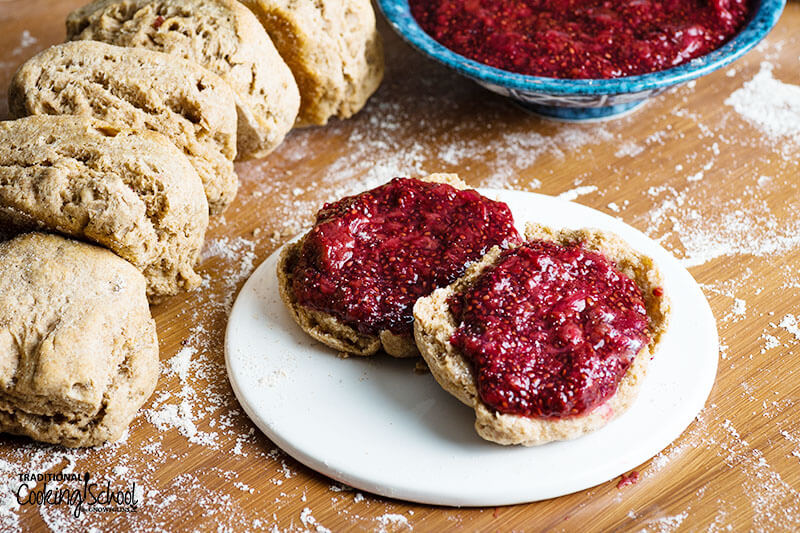 Image of sprouted spelt biscuits on a table with one biscuit cut open and topped with jam.