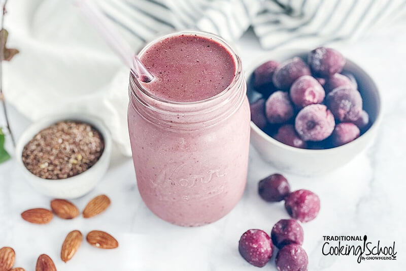 Pink-colored hormone balancing smoothie in a small glass jar with a glass straw, surrounded by cherries, flax seeds, and almonds.