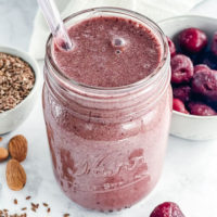 pink smoothie in a glass jar with a glass straw. Cherries, flax seeds and almonds are scattered in the background