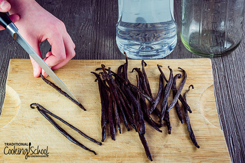 hand chopping vanilla beans in half lengthwise on a cutting board, with a bottle of vodka and an empty jar in the background