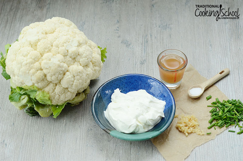 array of ingredients: one head of cauliflower, bowl of sour cream, apple cider vinegar, herbs, and spices