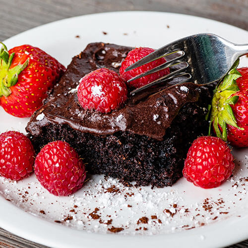 A white plate with a piece of chocolate frosted chocolate cake topped with fresh raspberries and strawberries
