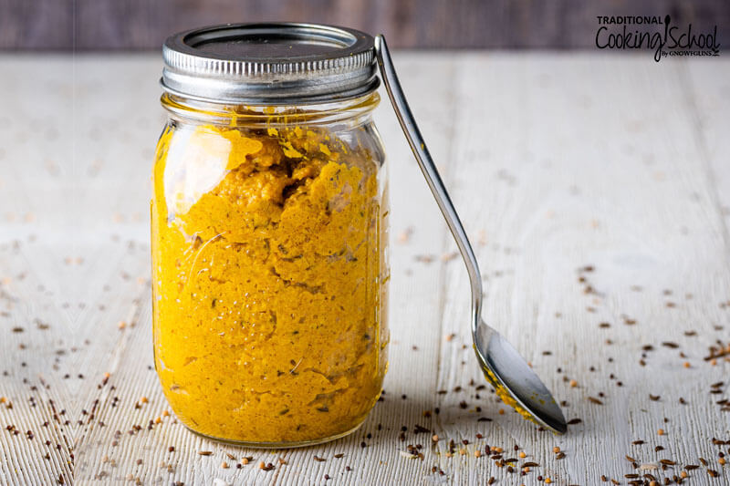 Homemade mustard in a mason jar with a spoon leaning up against the jar and mustard seeds spread on the counter.