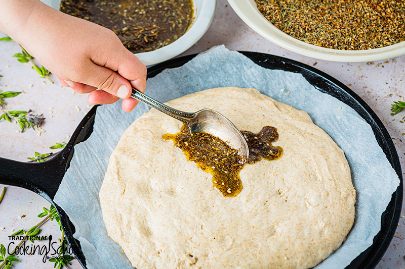 Manakish dough on a cast iron pizza pan being spread with za'atar and olive oil mixture with a spoon.