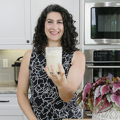 smiling woman in a kitchen holding out her small jar of sourdough starter