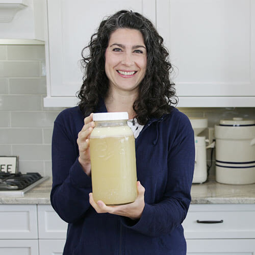 woman smiling in her kitchen holding forward a half-gallon jar of broth