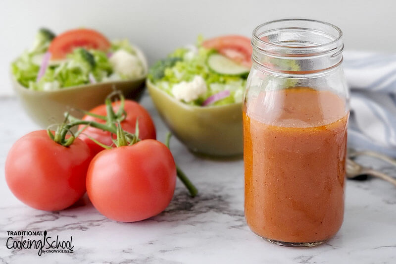 small glass jar of tomato salad dressing in front of fresh green salads and three tomatoes on the vine