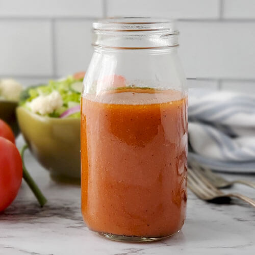 small glass jar of rust-colored tomato salad dressing with a fresh green salad in the background