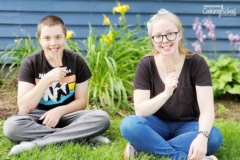 two teenagers sitting on the grass outside, smiling while holding up homemade fudge pops