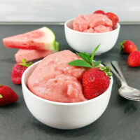two bowls of pink-colored sorbet garnished with fresh strawberries, with watermelon wedges stacked in the background