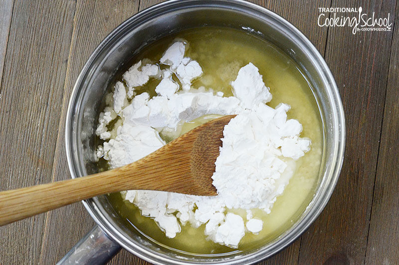 Wooden spoon stirring arrowroot powder into a saucepan of other ingredients for making deodorant.