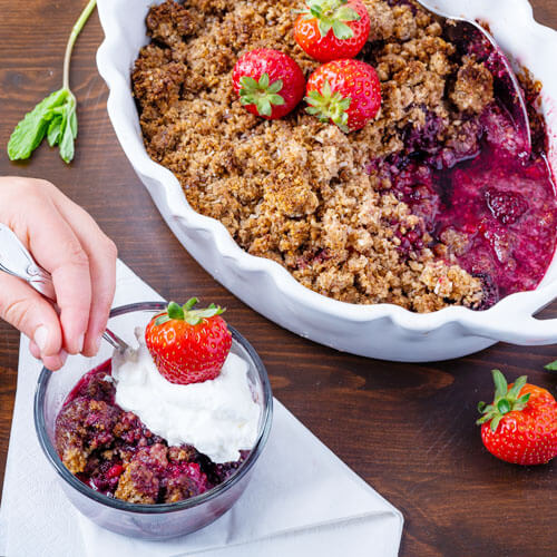 Small hand digging into a bowl of berry cobbler topped with whipped cream and a fresh strawberry, with the entire cobbler in the background.