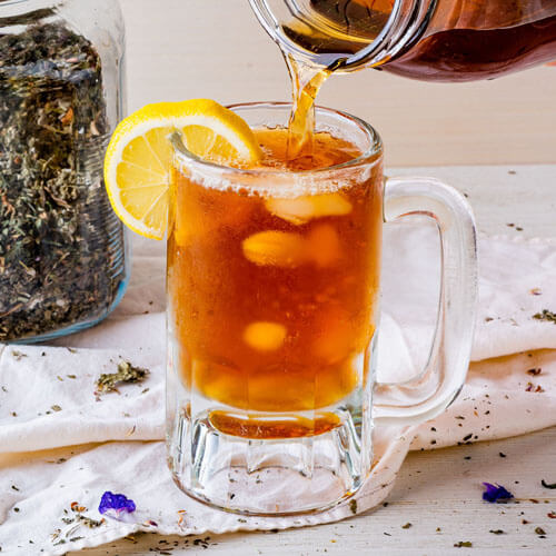 Pouring a cold herbal infusion into a glass to make iced tea. A lemon slice is stuck on the rim of the glass.