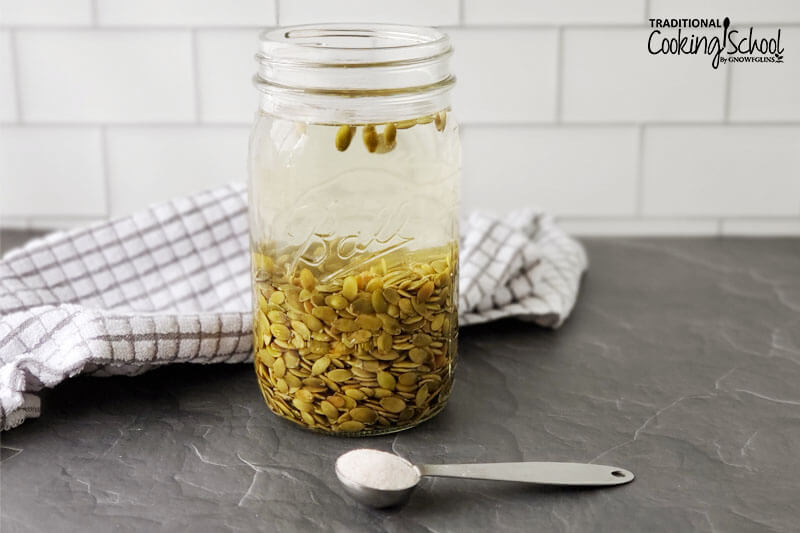 raw pumpkin seeds soaking in a glass jar with a teaspoon of salt in the foreground