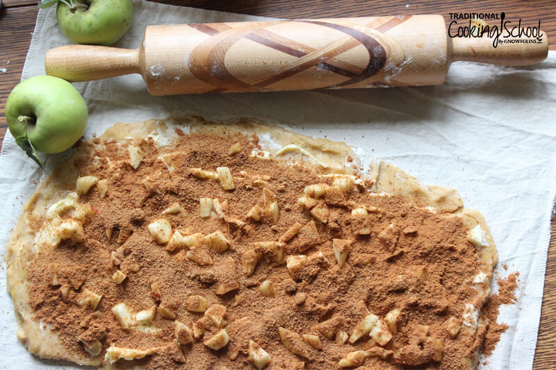 Cinnamon roll dough rolled out in a rectangle and sprinkled with spices, sugar, and apple slices. A rolling pin is nearby.