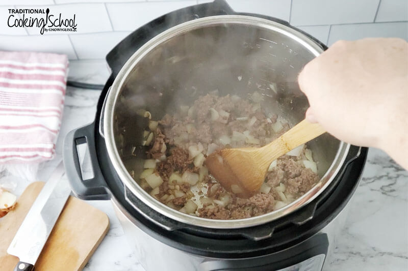 Woman's hand stirring ground meat with a wooden spoon as it browns in the Instant Pot.