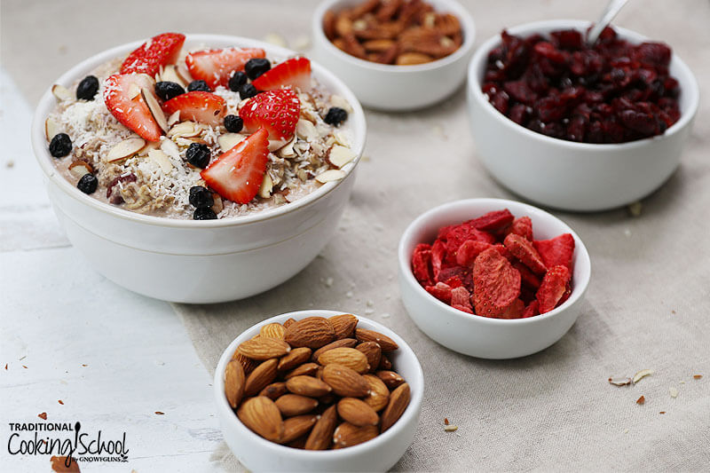 Bowl of muesli topped with fresh berries surrounded by small bowls of other ingredients and toppings: almonds, freeze-dried strawberries, dried cranberries, and pecans.