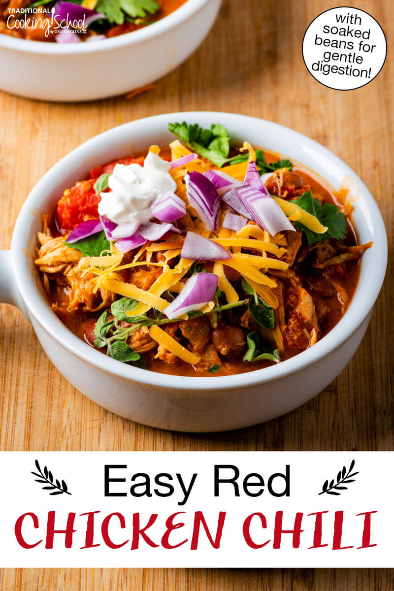 Bowl of chili topped with sour cream, red onion, cilantro, and grated cheddar cheese. Text overlay says: "Easy Red Chicken Chili (with soaked beans for gentle digestion!)"