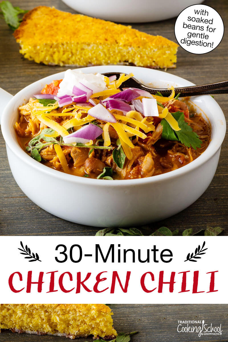 Bowl of chili topped with sour cream, red onion, cilantro, and grated cheddar cheese, with a slice of cornbread in the background. Text overlay says: "30-Minute Chicken Chili (with soaked beans for gentle digestion!)"