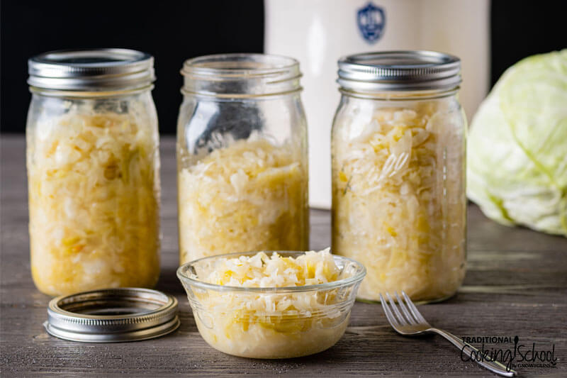 Homemade sauerkraut in a small glass bowl with jars of finished kraut in the background.