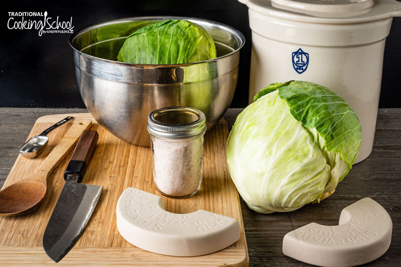 Ingredients and equipment needed for making sauerkraut: large mixing bowl, cabbage, sea salt, fermenting weights, stoneware crock, wooden spoon, measuring spoon, cutting board, and knife.