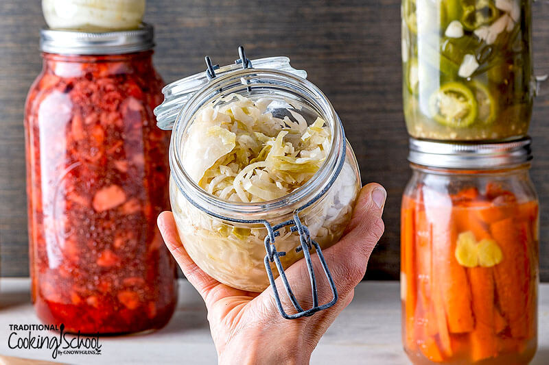 Person's hand holding up a glass jar of homemade sauerkraut with other ferments in the background.