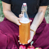 Woman holding up a swing-top glass bottle filled with foamy apple cider.