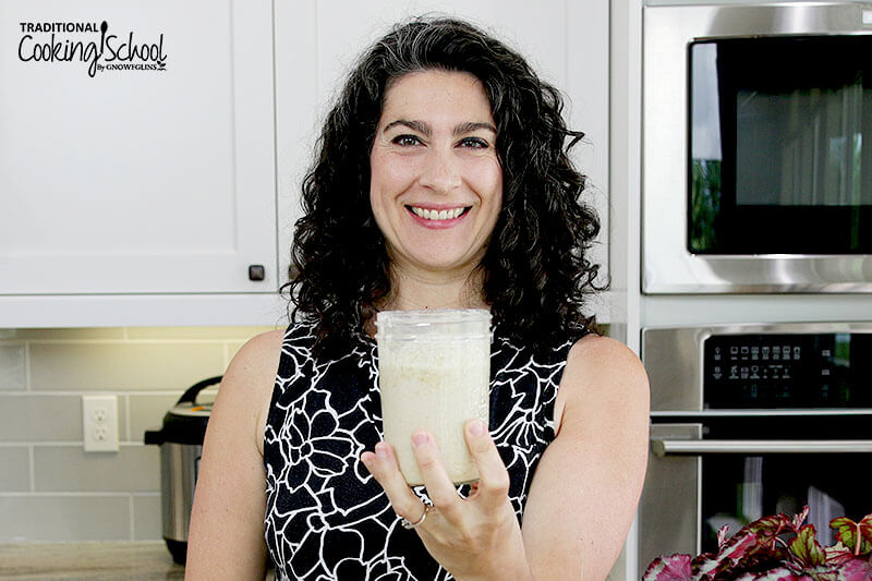Smiling woman in her kitchen holding up a jar of sourdough starter.
