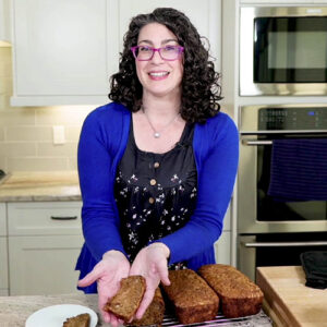 A smiling woman in her kitchen holding up a slice of freshly baked banana bread.