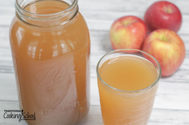 Photo of a half gallon jar and glass, both full of apple cider switchel with apples in the background.