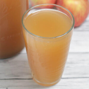 Photo of a glass of apple cider switchel with apples in the background.