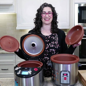 Photo of a smiling woman in her kitchen with two VitaClay cookers in front of her. She is holding a lid in each hand.